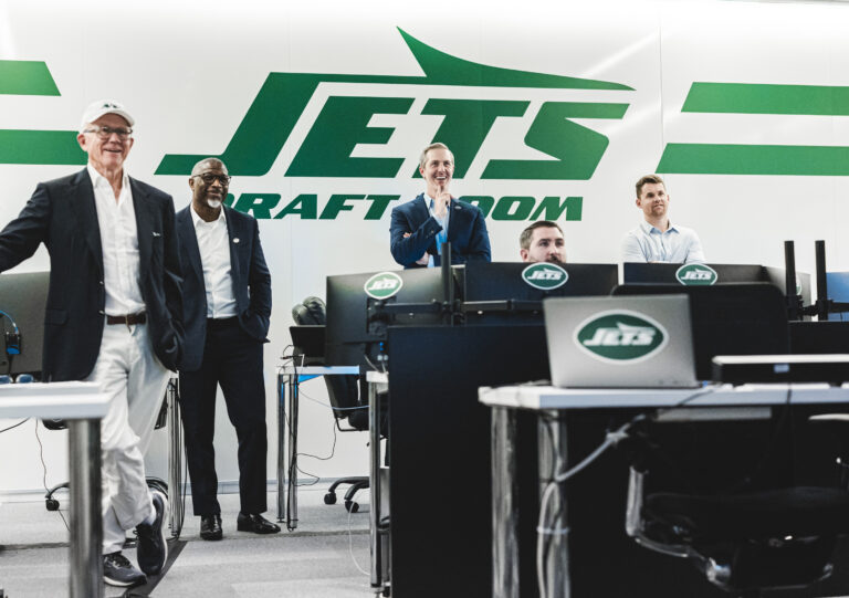 A group of men stand and sit in the NY Jets draft room.