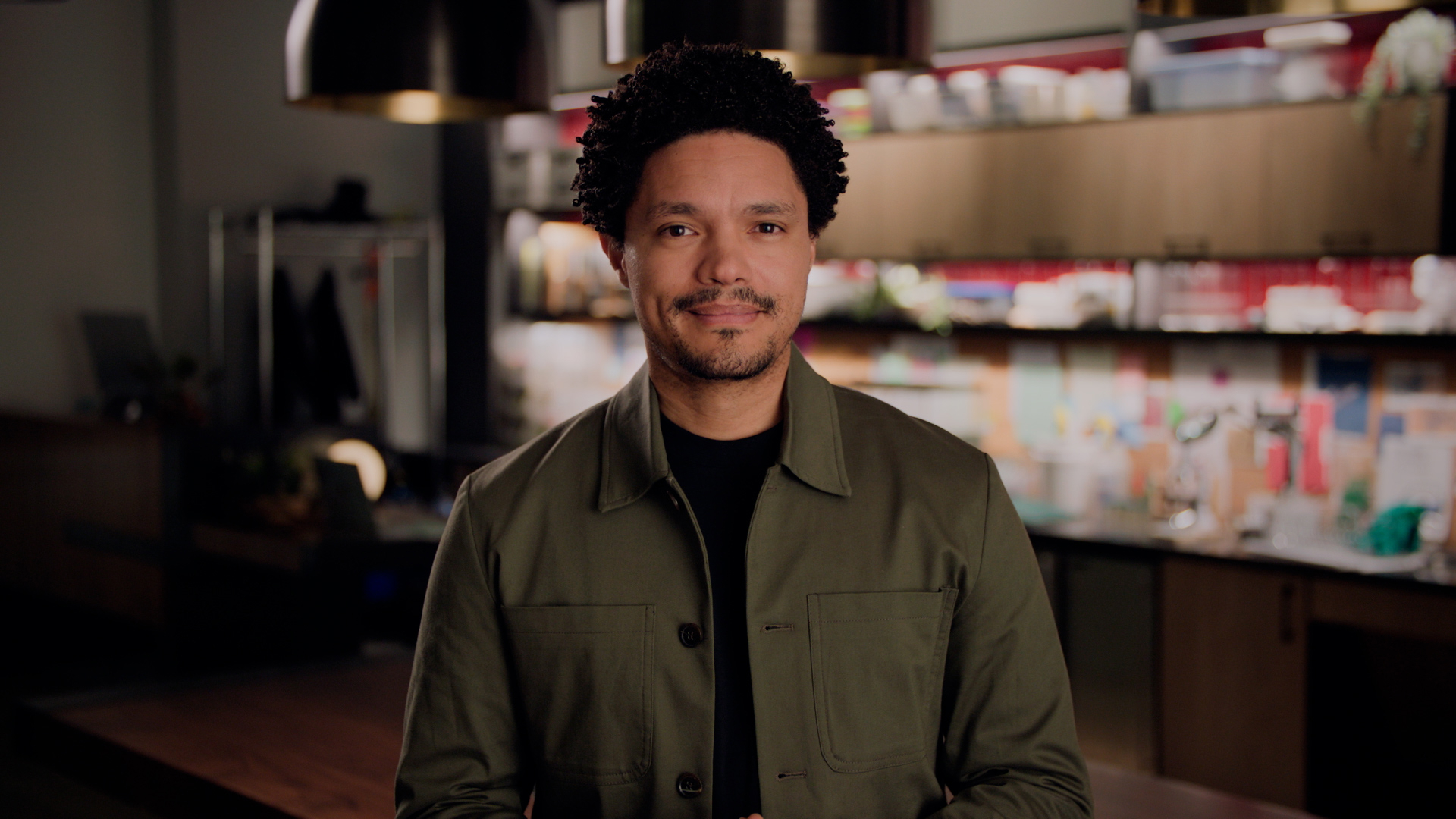 A man with curly hair stands in a kitchen, wearing a green jacket over a black shirt. The kitchen has wooden cabinets, shelves with various items, and is softly lit.