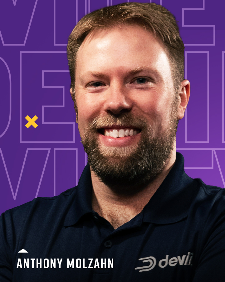 A man with a beard smiles at the camera, wearing a navy blue shirt with a "devii" logo. The background is purple with large, light text and a small yellow "x" shape.