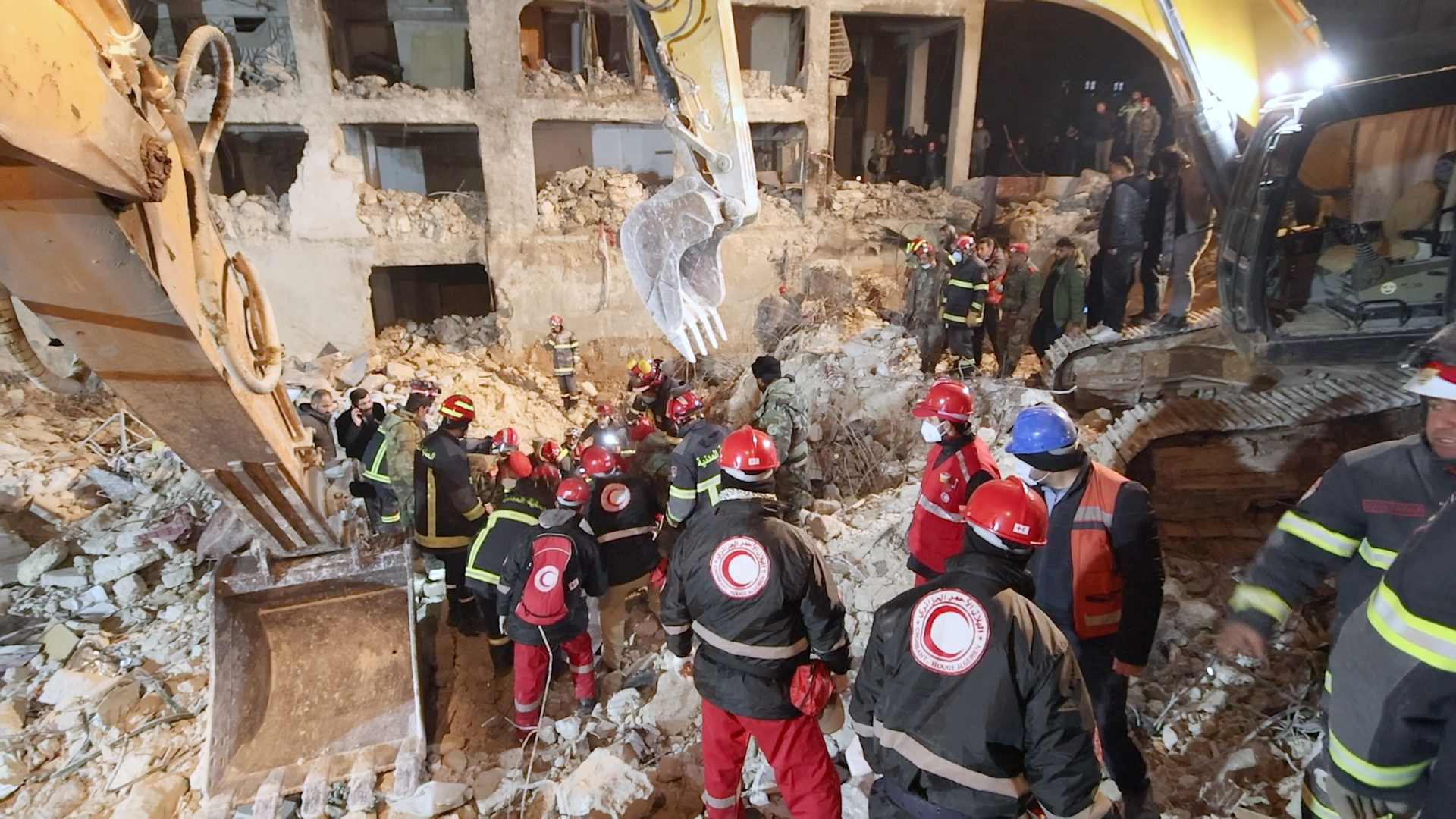 Rescue workers in helmets and uniforms search through rubble with excavators near a damaged multi-story building at night.